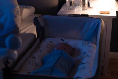 Koza kundaklama çantasına sarılmış bir bebek bebek bakıcısı kamerasıyla ebeveyn yatağının yanındaki bir karyolada huzur içinde uyuyor. Güvenli yeni doğan bebek uykusu. Modern teknoloji. Yumuşak seçici odak.