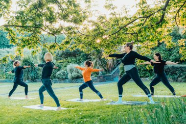 Park 'ta kadın yoga dersi. Bir grup farklı kadın esneme pozu veriyor yeşil çimlerin üzerinde eğitmenle birlikte egzersiz yapıyorlar. Sağlık, refah ve akıl sağlığı için spor faaliyetleri. Seçici odak.