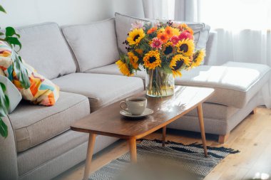 Bir fincan kahve ve vazo, üzerinde kocaman çok renkli bir çiçek buketi, modern, rahat oturma odasında, gri kanepeli sehpanın üzerinde. Açık uzay ev iç tasarımı. Konforlu Tasarım ev dekorasyonu