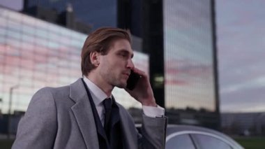 Zarif bir iş adamının portresi bir iş arabasının yanında duruyor ve bir cep telefonuyla konuşuyor, takım elbiseli üst düzey bir yönetici, akşam vakti, gün batımı.