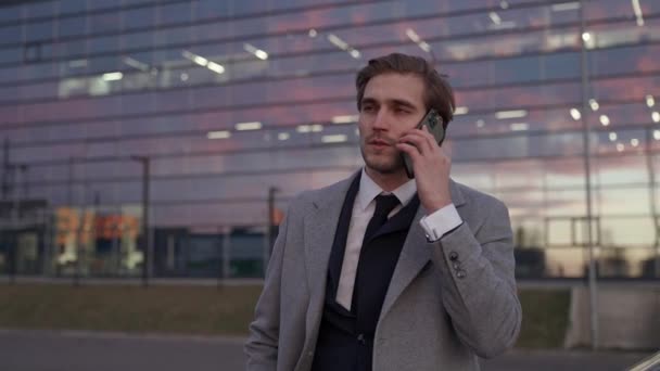 一个年轻商人站在一辆商用车旁 用手机交谈 身穿西服的高级经理 傍晚时分 身后是摩天大楼的画像 — 图库视频影像