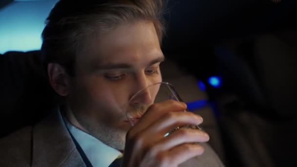移動中の車に座ってウイスキーを飲んでいる若いビジネスマンの肖像画 スーツのトップマネージャーはビジネス車の後ろに座っています ナイトライド — ストック動画
