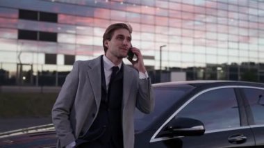 El bilgisayarı, zarif bir iş adamının portresi bir iş arabasının yanında duruyor ve cep telefonuyla konuşuyor, takım elbise giymiş üst düzey bir yönetici, akşam vakti, gün batımı..