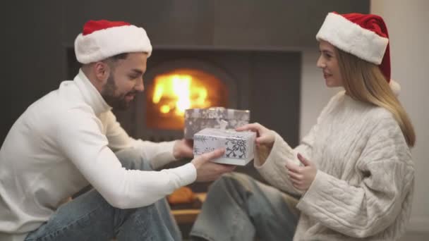 クリスマスを祝い 陽気なカップルはお互いに贈り物を持って箱を与え サンタ帽子の男女は暖炉に座ってシャンパン 冬の休日を飲みます — ストック動画