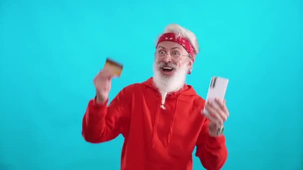 头发灰白 头戴红色帽衫的嬉皮士男人通过智能手机和信用卡成功地在网上购物 他是一个穿着蓝色背景的时髦白人男人 — 图库视频影像