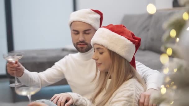クリスマスを祝い 若いカップルはリビングルームの床に座り サンタハットでシャンパン 男と女性を飲んで 冬の休日 — ストック動画