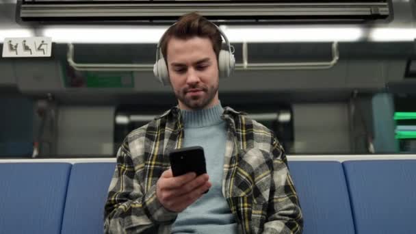 地下に集中した男性が地下鉄に乗り ワイヤレスヘッドフォンで音楽を聴き スマートフォン 公共交通機関を使用する — ストック動画
