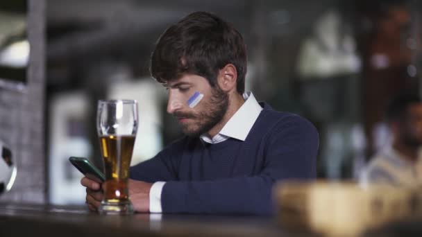 サッカーファンはサッカーの試合中にバーに座っていて スマートフォンを使用して 男は携帯電話を使ってテキストを読んでいます — ストック動画