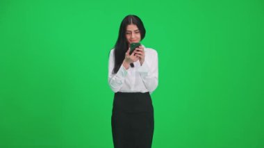 Takım elbiseli neşeli bir kadın krom anahtar arka planda duruyor, akıllı telefon kullanıyor ve ekrana bakıyor, internette sörf yapıyor, yeşil arka plan, kromakey şablonu.