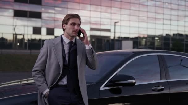 ビジネスカーの近くに立って 携帯電話で話す若いビジネスマン スーツのトップマネージャー バックグラウンドの高層ビルで — ストック動画