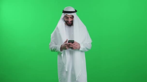 一名阿拉伯男子身穿绿色背景的白色长袍 头戴坎杜拉式便服 手持智能手机 在网上冲浪 带着铬钥匙模板 — 图库视频影像