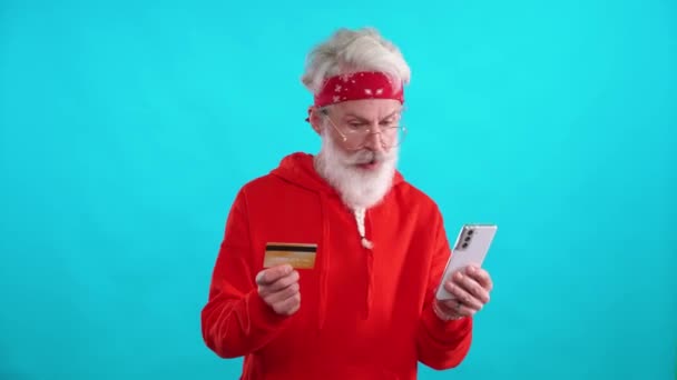 头戴红色帽衫的白发男士通过智能手机和信用卡在网上成功购物 他是一位穿着蓝色背景的时髦的高加索男士 — 图库视频影像