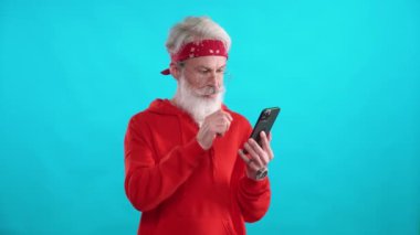 Kırmızı kapüşonlu yetişkin gri saçlı adam internette iyi ürünler ve yararlı bilgiler buluyor. Akıllı telefon kullanıyor. Mavi arka planda şık bir beyaz adam..
