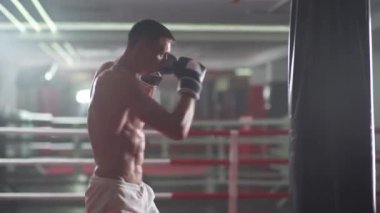 Erkek dövüşçü yumruklarını eğitir, kum torbasını döver, boks salonunda antrenman yapar, elleriyle ve ayaklarıyla saldırır..