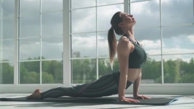 Genç bir kadın ışıkla dolu beyaz bir odada yoga yapıyor, kız yoga yapıyor ve büyük pencerenin yanında elementler sergiliyor..