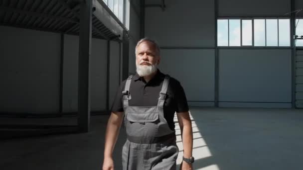 大人の灰色の髪の男性労働者は倉庫を歩き 作業エンジニアは空の物流倉庫 映画の照明の領域を歩きます — ストック動画