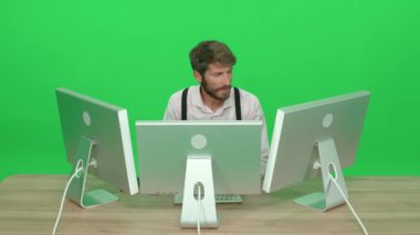 Masada otururken bilgisayar üzerinde çalışan odaklanmış yazılım geliştiricisi, yeşil arkaplan, monitörlerin arkasında çalışan bir adam, kroma anahtar şablonu, arka görünüm.
