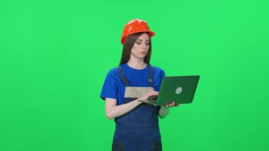 Üniformalı ve koruyucu başlıklı odaklanmış bir kadın işçinin portresi, mühendis ayağa kalkar ve yeşil arka planda dizüstü bilgisayar kullanır, kromakey üzerinde kadın mühendis..