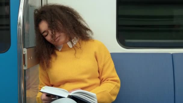 地下鉄で眠っているダークスキーの女性が手元に本を持って 若い学生が大学 公共交通機関で勉強する — ストック動画