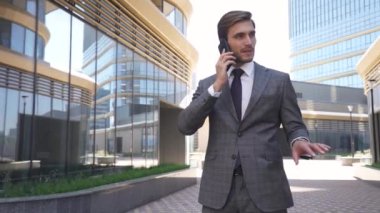 Takım elbiseli genç işadamı cam bir gökdelenin yanında duruyor ve cep telefonuyla konuşuyor, modern ofisin en üst düzey yöneticisi..
