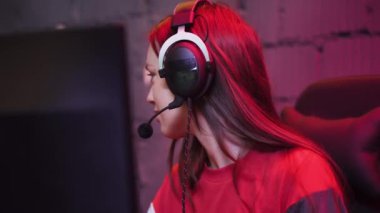 Kulaklıklı genç kız oyuncu, video oyunu oynuyor, klavyede daktilo kullanıyor, mercan rengi ve neonu aydınlatıyor, oyunda siber sporcu, oyuncular arasında iletişim kuruyor..
