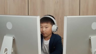 Asyalı genç bir çocuk masada otururken bilgisayar üzerinde çalışıyor, bir çocuk monitörlerin arkasında çalışıyor ve çalışıyor..