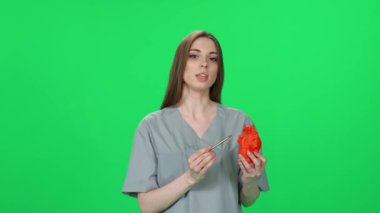 Kadın doktor portresi, güzel kadın kameraya bakar ve yeşil arka planda insan kalbinin plastik maketini kullanarak kalbin yapısını kelimelerle betimler..