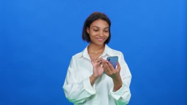 İnternetten alışveriş yapan, beyaz gömlekli koyu tenli bir kadın iyi ürünler buluyor ve akıllı telefon kullanıyor. Mavi arka planda şık bir Afrikalı kadın..