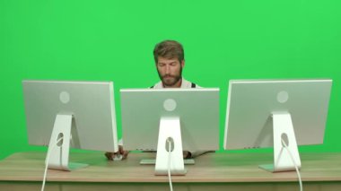 Masada otururken bilgisayar üzerinde çalışan odaklanmış yazılım geliştiricisi, yeşil arka plan, monitörlerin arkasında çalışan bir adam, kroma anahtar şablonu.