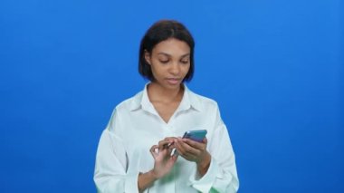 İnternetten alışveriş yapan, beyaz tişörtlü, duygusal esmer bir kadın iyi ürünler buluyor ve akıllı telefon kullanıyor. Mavi arka planda şık bir Afrikalı kadın..
