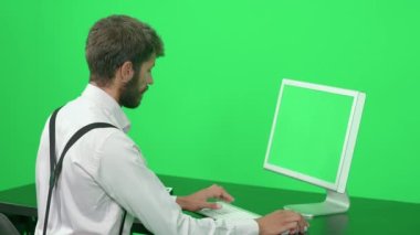 Masada otururken bir bilgisayar üzerinde çalışan odaklanmış yazılım geliştiricisi, yeşil arkaplan, krom anahtar şablonu, arka görünüm.