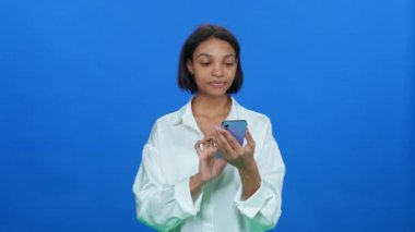 İnternetten alışveriş yapan, beyaz tişörtlü, duygusal esmer bir kadın iyi ürünler buluyor ve akıllı telefon kullanıyor. Mavi arka planda şık bir Afrikalı kadın..