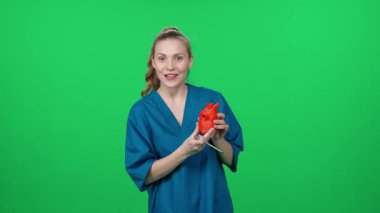 Kardiyolog, kadın doktor portresi, güzel kadın kameraya bakar ve yeşil arka planda insan kalbinin 3 boyutlu plastik maketini kullanarak kalbin yapısını gösterir..