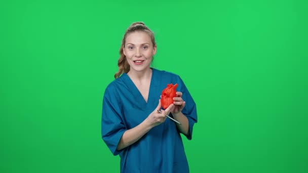 婦人科医 女性医師の肖像画 かわいい女性はカメラを見て 緑色の背景に人間の心臓の3Dプリントされたプラスチックのモックアップを使用して心臓の構造を示します — ストック動画