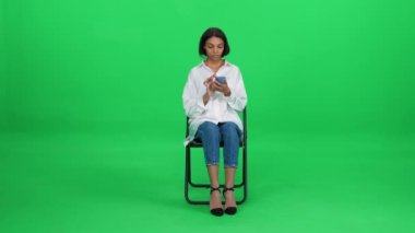 Beyaz gömlekli şık, koyu tenli bir kadın ofis sandalyesinde oturuyor ve akıllı telefon kullanıyor, yeşil arka plan, bekleme süreci..