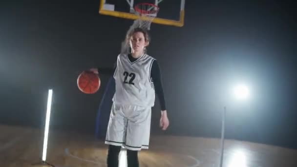 年轻女篮球运动员的形象在赛前把球打出地板 专业篮球运动员看着镜头 室内操场 — 图库视频影像