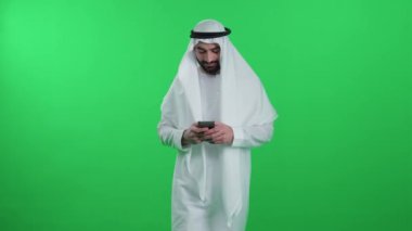 Yeşil arka planda beyaz kanduralı bir Arap, akıllı telefon kullanarak yürüyor ve internette geziniyor, kromakey şablonu.