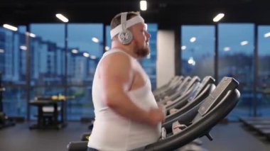 Motivasyon ve kendini geliştirme, koşu bandında koşan ve kulaklıkla müzik dinleyen şişman adam, spor salonunda aerobik egzersiz ve dayanıklılık eğitimi..