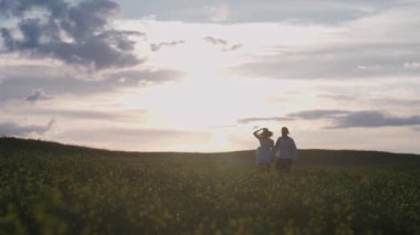 Kırsal kesimde, neşeli bir çift doğada dinleniyor, bir erkek ve bir kadın kolza tohumu tarlasında koşuyorlar, el ele tutuşuyorlar, gün batımı, 4k ağır çekim.