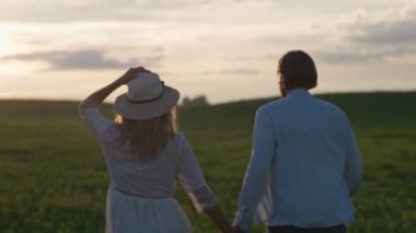 Kırsal kesimde gün batımı, doğada genç bir çift, bir erkek ve bir kadın kolza tohumu tarlasında koşuyorlar, el ele, arka manzara, 4k yavaş çekim.