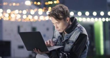 Genç adam şehir merkezinde, modern bir yerleşim bölgesinde oturan bir dizüstü bilgisayar kullanıyor. Arka planda bulanık ışıklar, 4K ağır çekim..