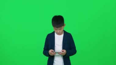 Asyalı okul çocuğu yeşil arka planda duruyor ve akıllı bir telefonla oyun oynuyor, çocuk zafere seviniyor, kromatonla şablon çiziyor..
