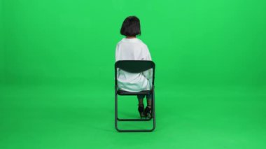 Beyaz gömlekli, zarif, koyu tenli bir kadın ofis sandalyesinde oturuyor, yeşil arka plan, bekleme süreci, arka görüş..