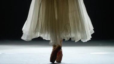 Beyaz etekli zarif bir kadın, bale yapıyor ve Pointe ayakkabılarıyla dengede duruyor, bacakları görüyor, koyu arkaplan, dikkatli adımlar atıyor..