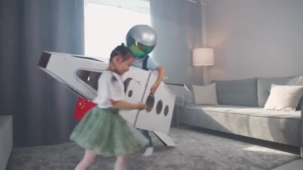 中国の子供たちは家のリビングルームで遊び 宇宙飛行士のヘルメットの男の子はダンボールのスペースシャトルで遊び 部屋の周りの弟の後を走るかわいい少女の妹 — ストック動画