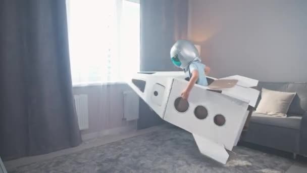 宇宙飛行士のコスチュームのアジアの少年は ダンボールのスペースシャトルで遊びます 夢想家は家で部屋の周りに実行します — ストック動画