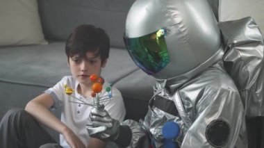 Küçük çocuk evdeki oturma odasında oynuyor, astronot kostümü giymiş bir baba oğluyla yerde oturuyor, çocuk güneş sisteminin oyuncak modeliyle oynuyor, astronomi okuyor..