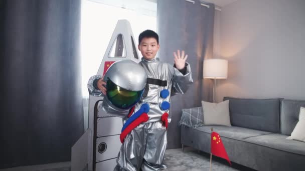 宇宙飛行士のコスチュームのアジアの十代の少年は 宇宙船のボール紙モデルの近くに立って ヘルメットを置き カメラと波を見ます 少年はリビングルームで宇宙飛行士を演じます — ストック動画