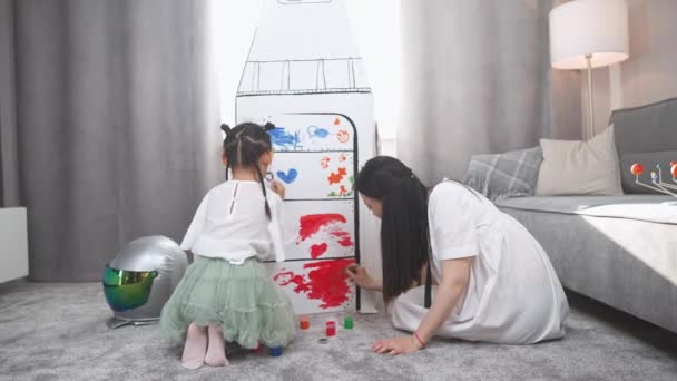 彼女の娘とアジアの女性は 家のリビングルームで遊び 床に座って 絵のスペースシップのボール紙モデルに母親と一緒に絵を描く — ストック動画