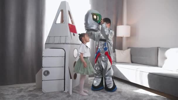 アジアの子供たちは家のリビングルームで遊びます 宇宙飛行士のコスチュームの男の子は ダンボールスペースシャトルの近くに彼女の妹と遊びます かわいい少女の妹は 宇宙飛行士のヘルメットに置きます — ストック動画